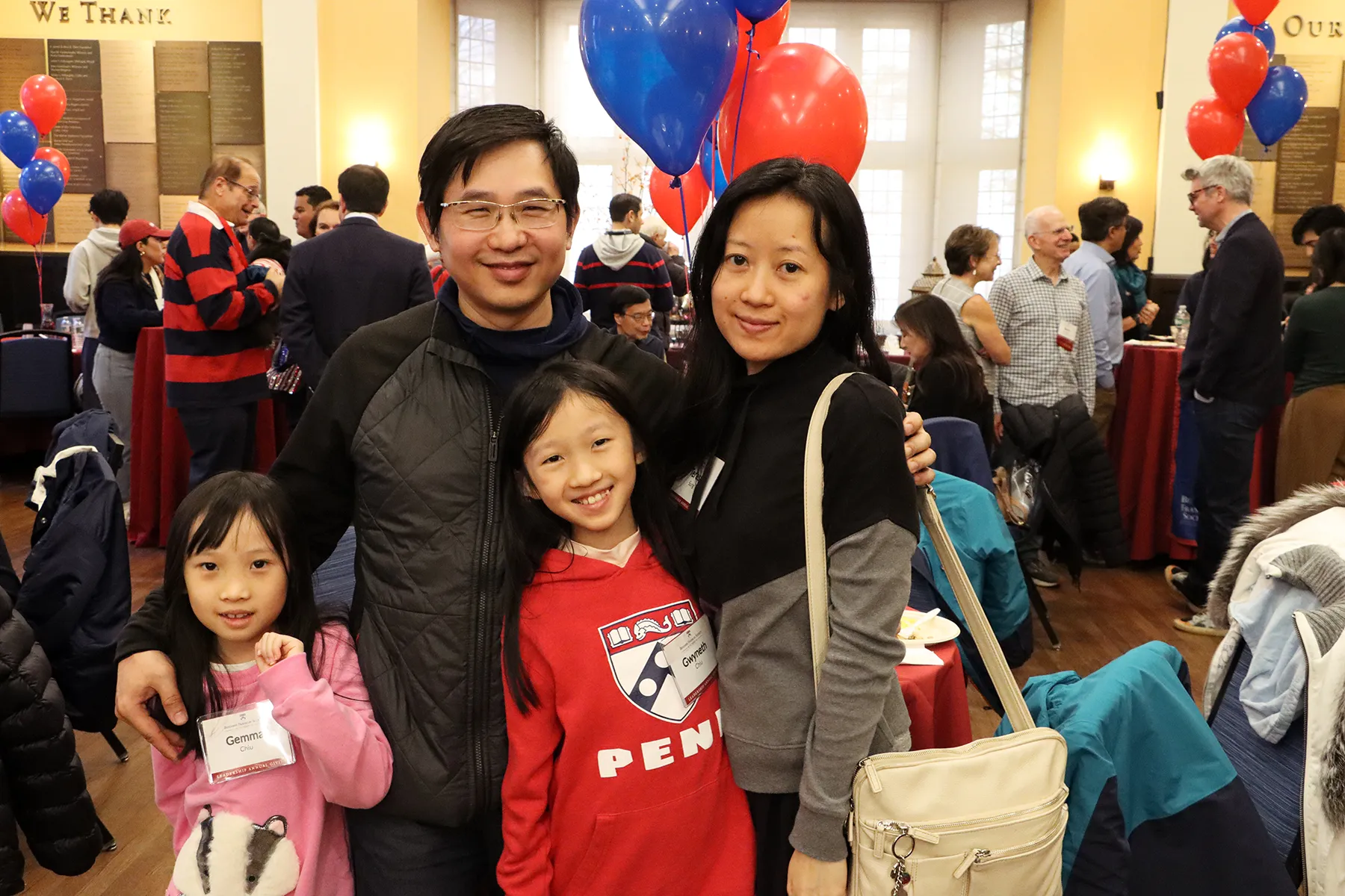 A family poses together at a University of Pennsylvania homecoming event indoors, with red and blue balloons and guests mingling in the background.