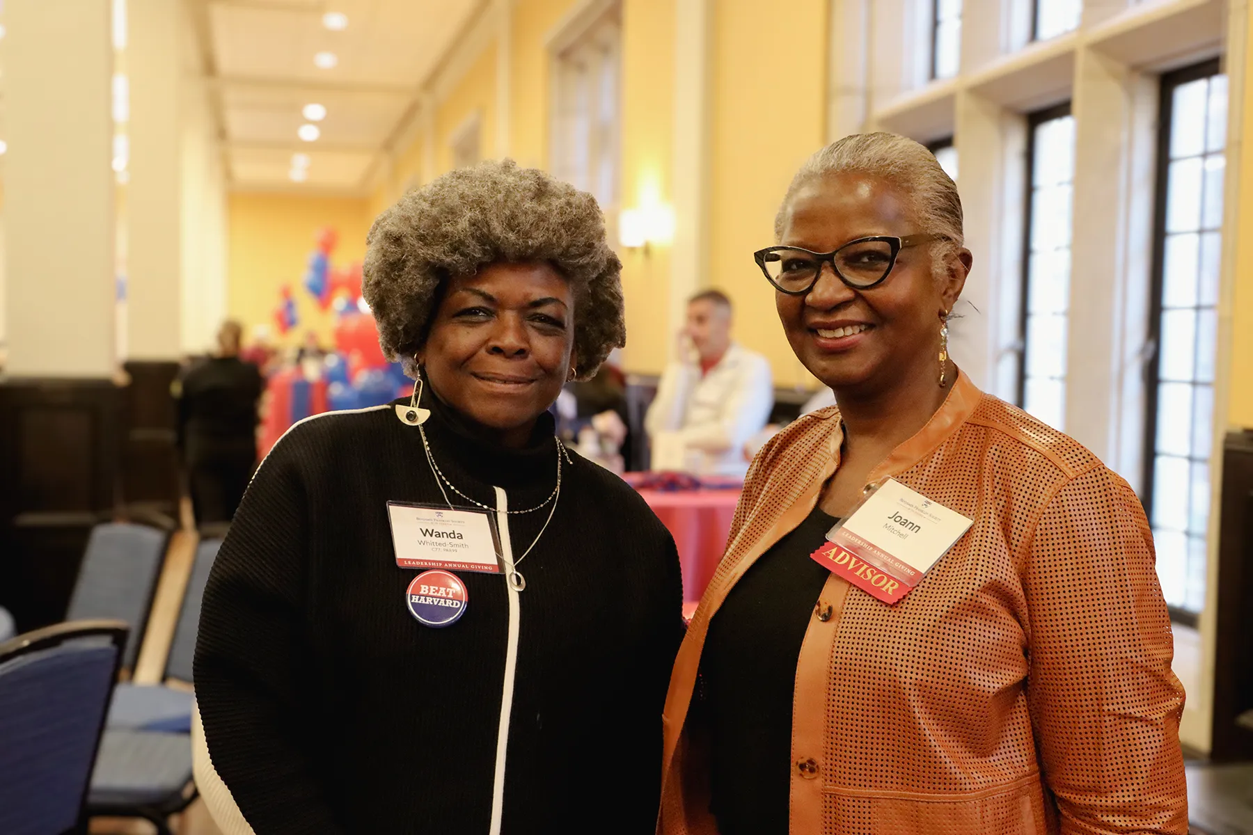 Two women smile for a photo at a University of Pennsylvania event, wearing name tags and standing in a warmly lit room decorated with balloons.