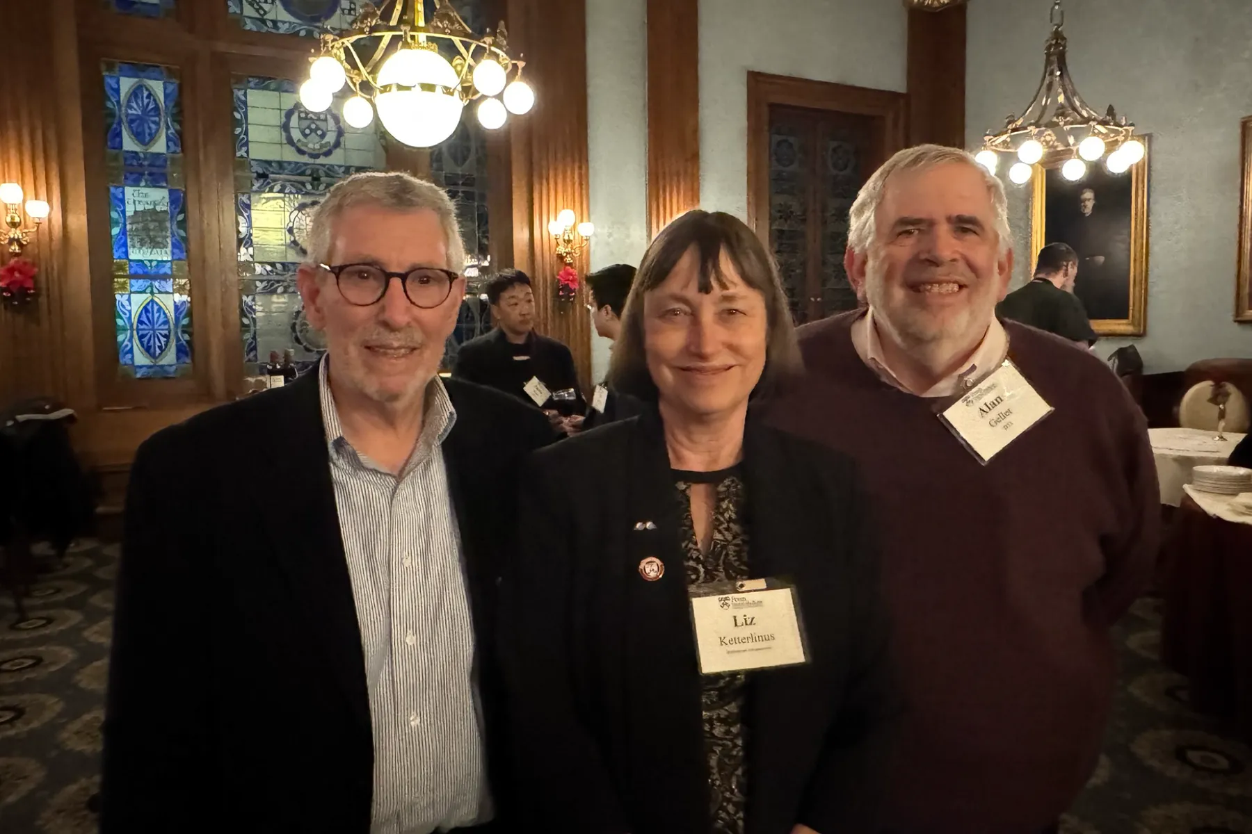 Three smiling guests at a University of Pennsylvania alumni event pose in front of stained glass windows and chandeliers in a warmly lit historic venue.