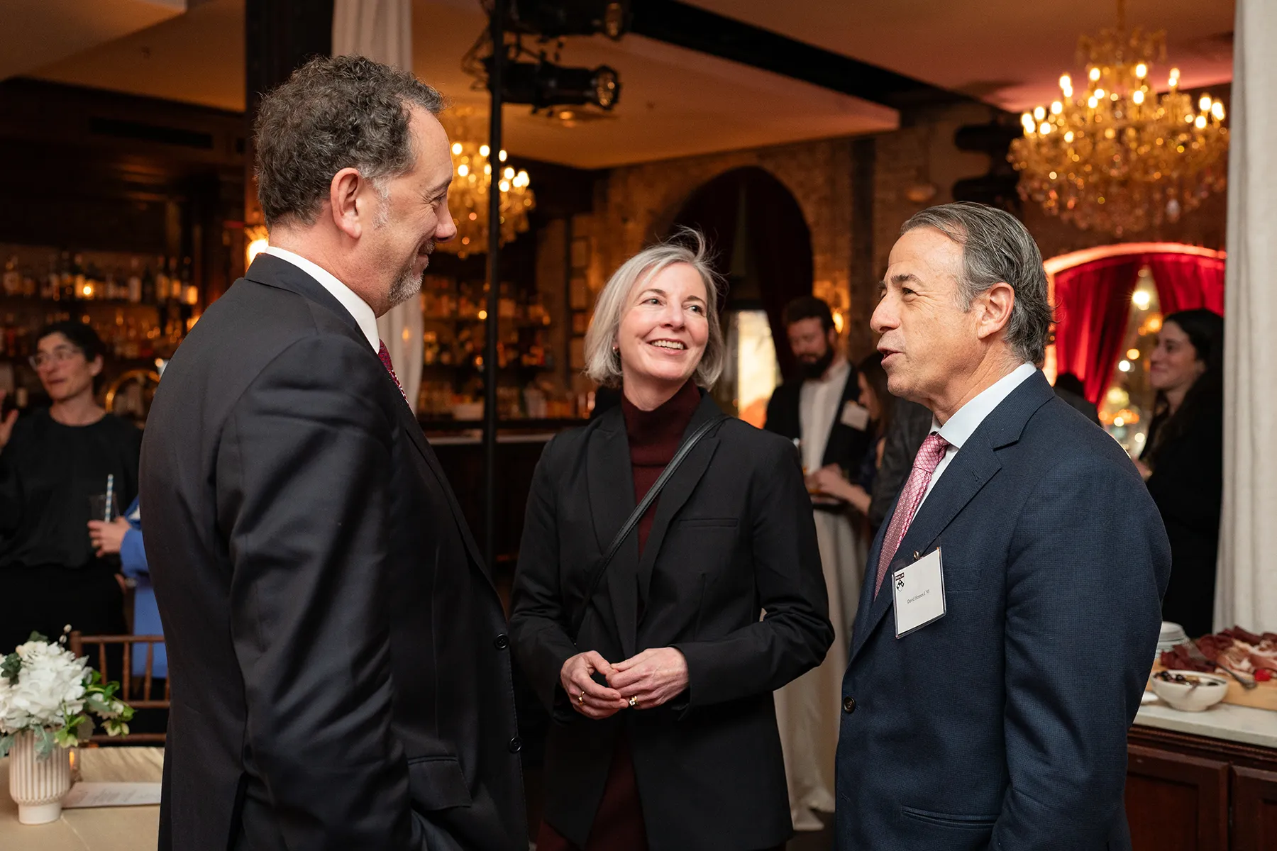 Three guests engage in friendly conversation at a University of Pennsylvania evening reception, standing near a bar in a warmly lit venue with chandeliers.