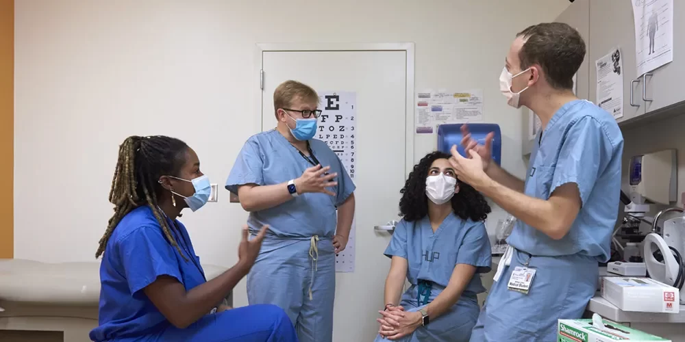 Four medical students wearing scrubs and masks having a discussion in a Penn Medicine exam room.