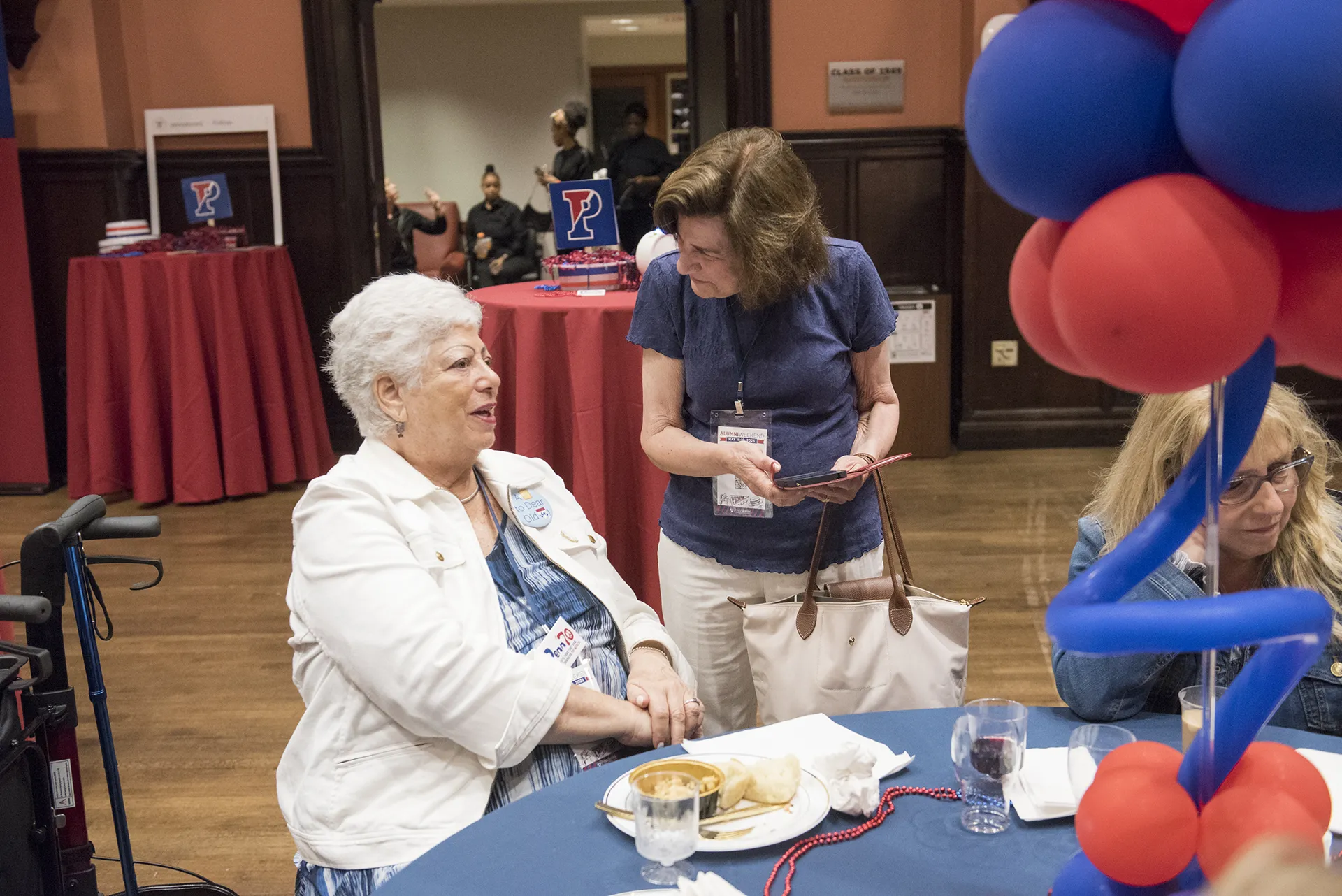 Two University of Pennsylvania Class of 1965 alumni warmly chatting at a decorated reunion table with red and blue balloon centerpiece