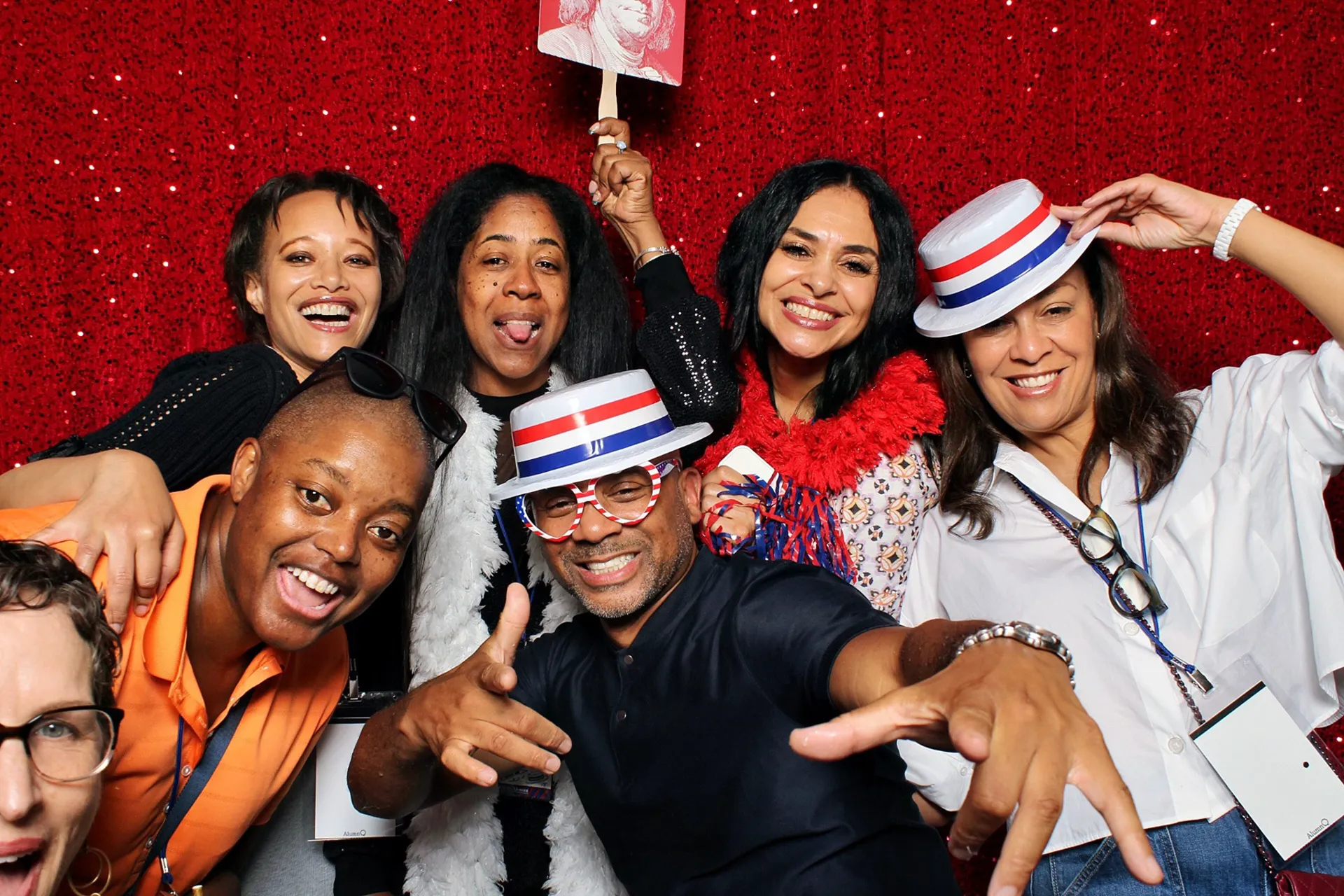 Smiling University of Pennsylvania Class of 1990 alumni wearing red-white-blue felt hats and festive props against a red sequined photo-booth backdrop at their 35th reunion