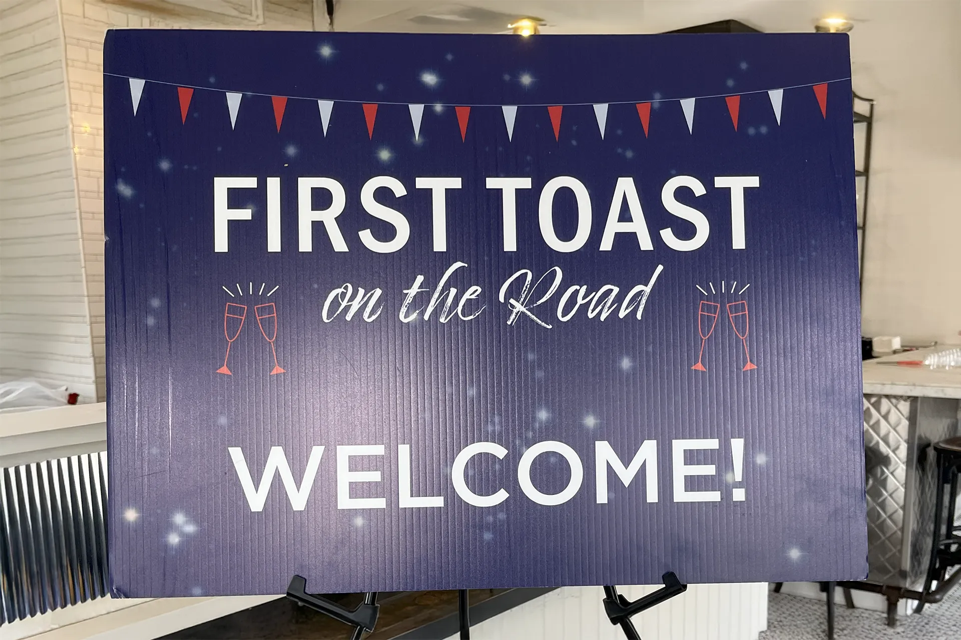A navy-blue welcome sign on an easel reading “FIRST TOAST on the Road WELCOME!” with festive bunting and champagne glasses graphics.
