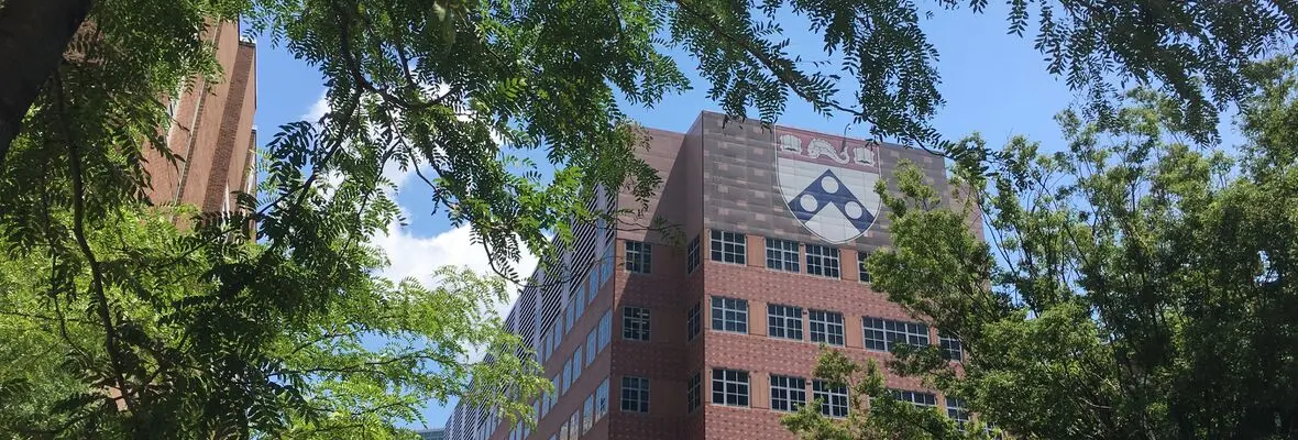 A red-brick Penn Medicine building is shown behind tree branches with the university emblem in view.