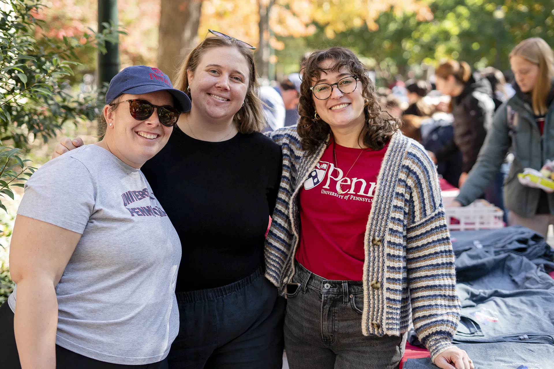 Three people smiling together at an outdoor Penn event with T-shirts on a table nearby.