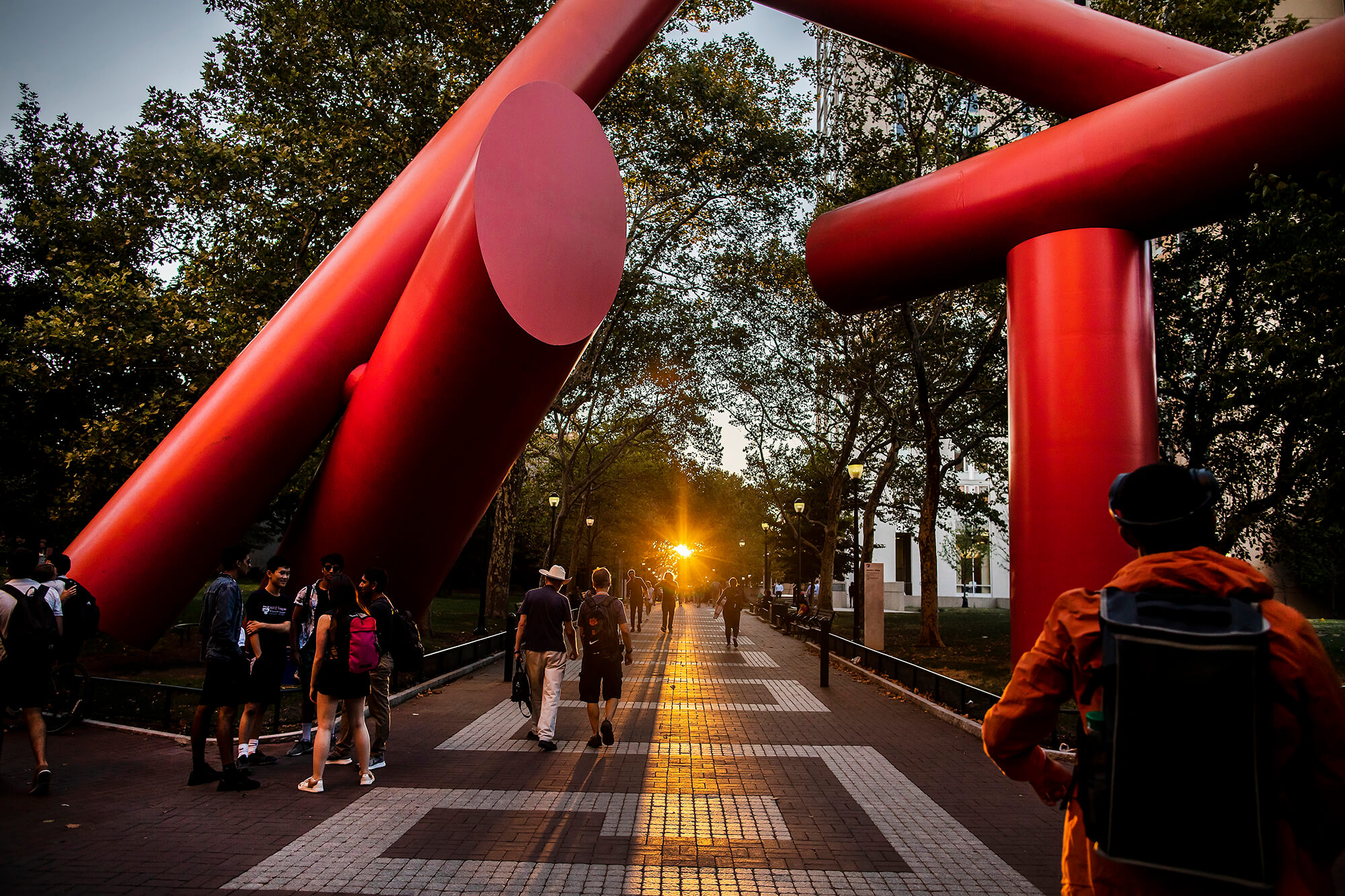 Students and other pedestrians walking along Locust Walk in Hamilton Village on the University of Pennsylvania campus at sunset, framed by the large red Covenant sculpture.