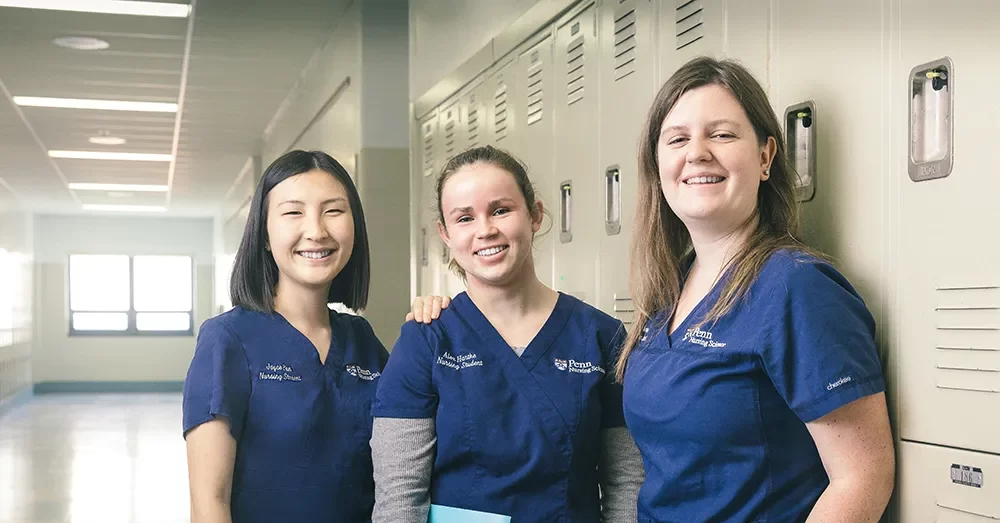 Three Penn Nursing graduate students in scrubs, smiling together in a hallway lined with lockers.