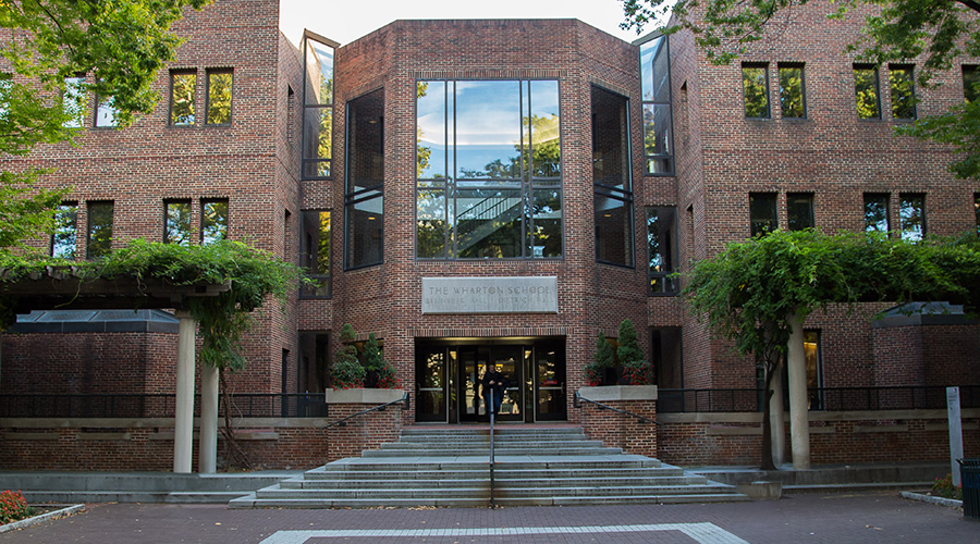 The Setinberg-Dietrich building on the University of Pennsylvania campus, showcasing a modern architectural style with red brick walls and large glass windows. The entrance is framed by columns of greenery, and the name 'THE WHARTON SCHOOL' is prominently displayed above the central glass doors.