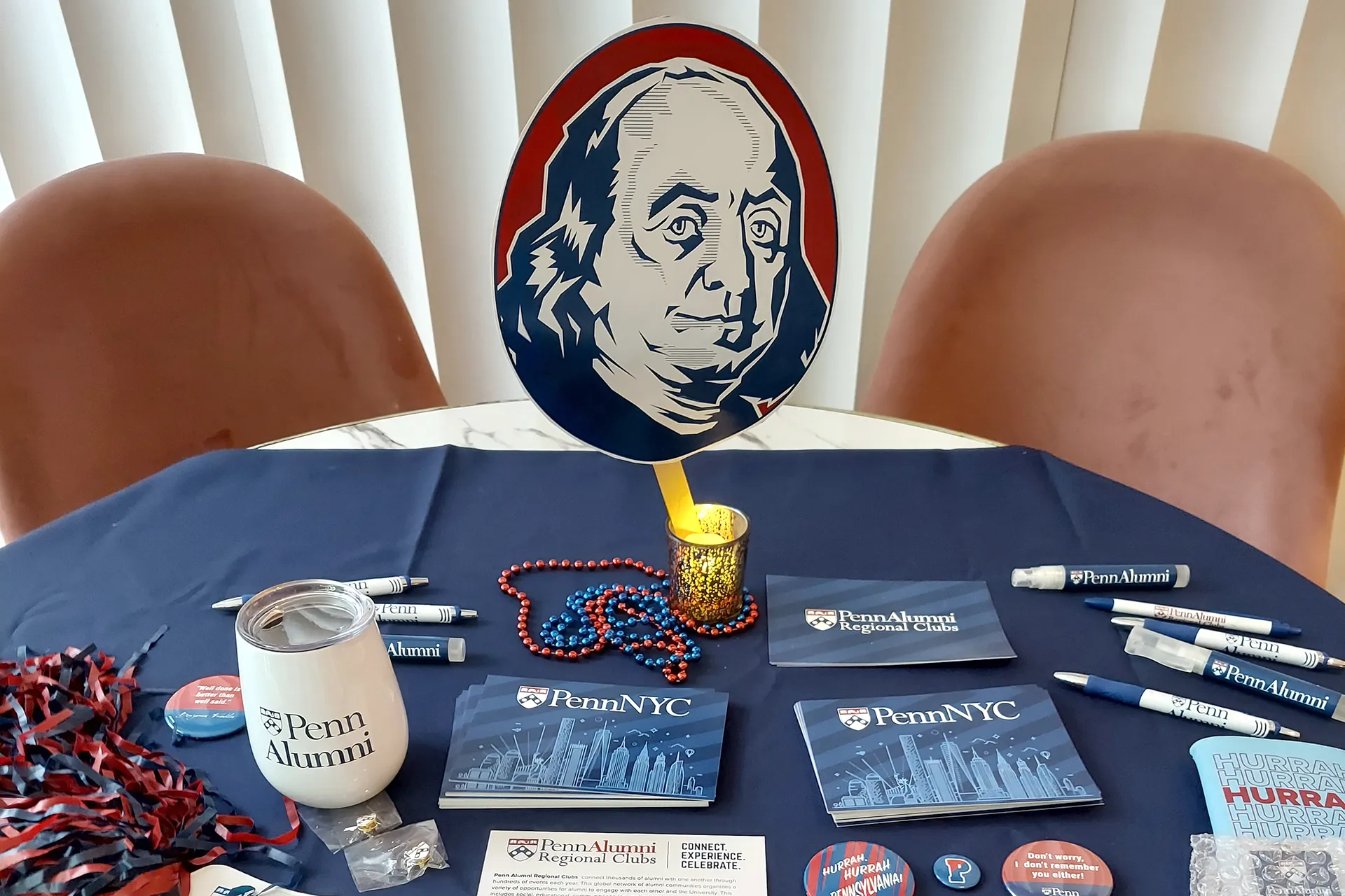 A table at an alumni event covered in a navy cloth, displaying Penn Alumni Regional Clubs brochures, PennNYC postcards, branded pens, lip balm, beads, buttons, a Penn Alumni tumbler, and a Benjamin Franklin cutout centerpiece.