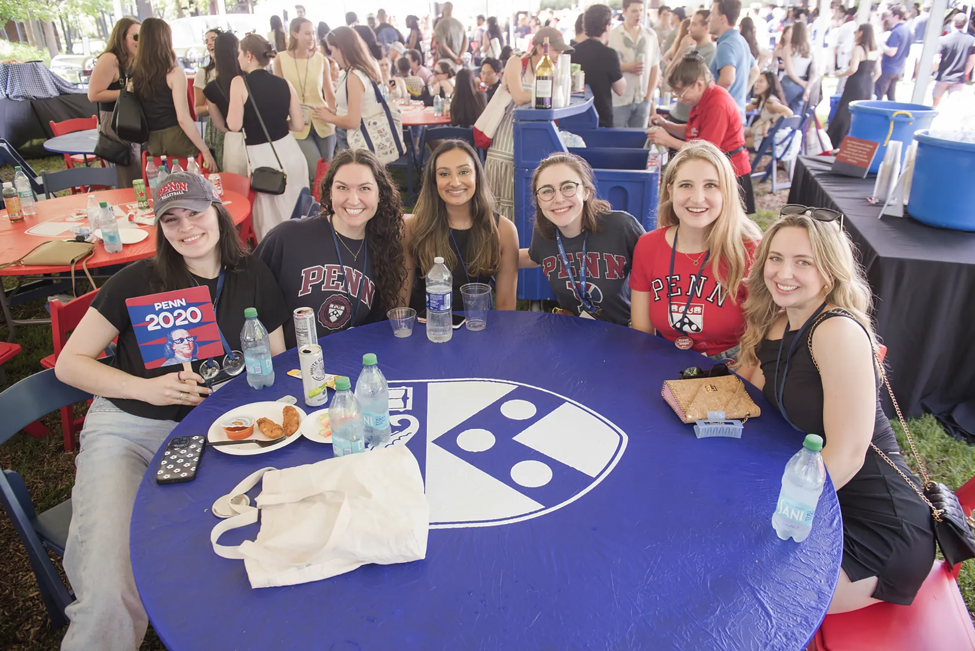 Six smiling alumni seated around a blue table with the Penn shield under an event tent, holding a “PENN 2020” fan sign, bottled water, and snacks.