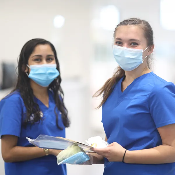 Two dental students wearing blue scrubs and face masks stand in a bright medical setting.