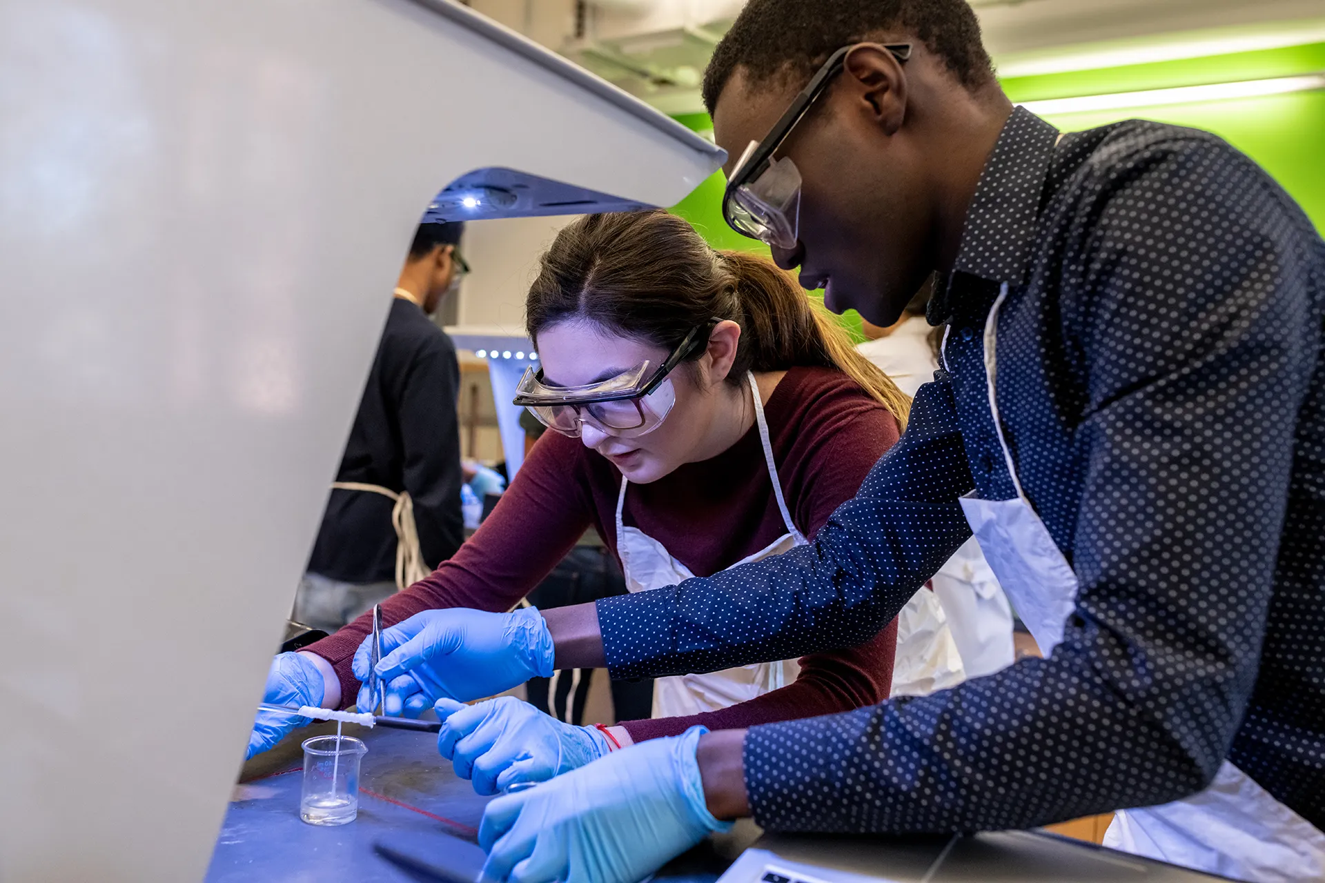 Two Penn students wearing gloves and goggles work together on a lab experiment, representing hands-on learning and scientific collaboration.