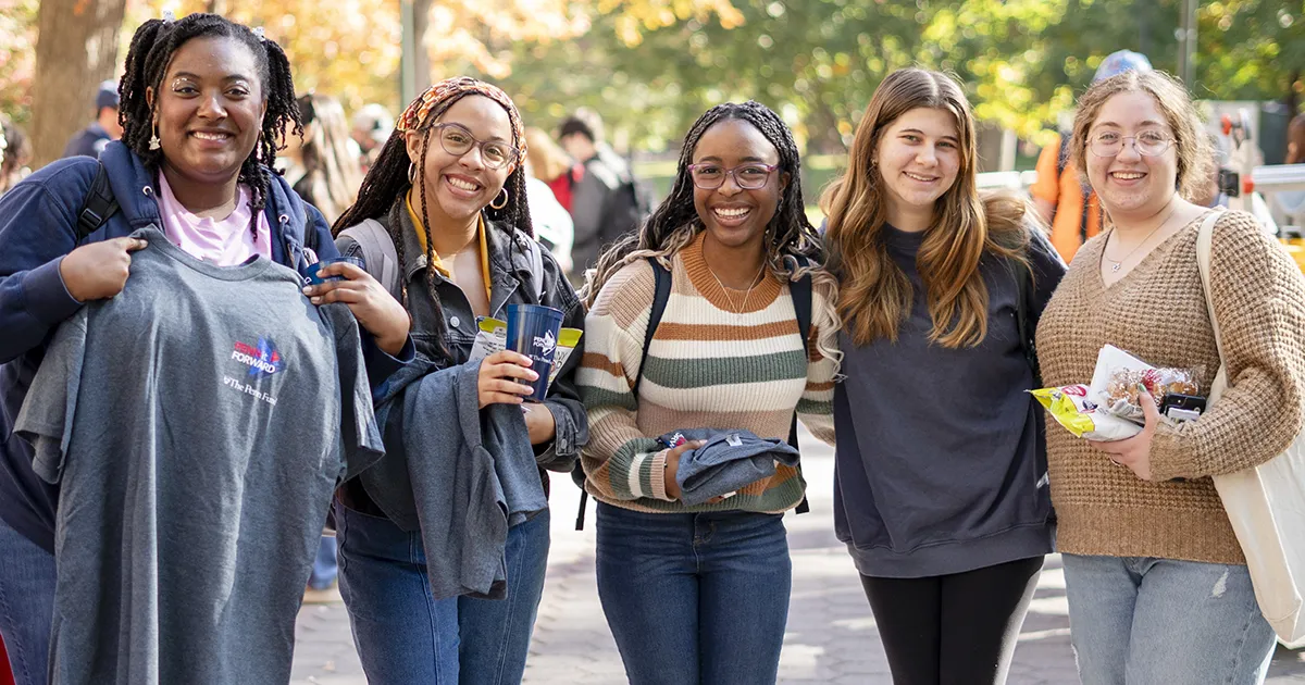 A group of Penn students smiling on Locust Walk, holding giveaways and t-shirts at an outdoor campus event.