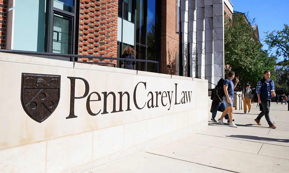 Students walking past a sign for Penn Carey Law on a bright day outside the law school building.