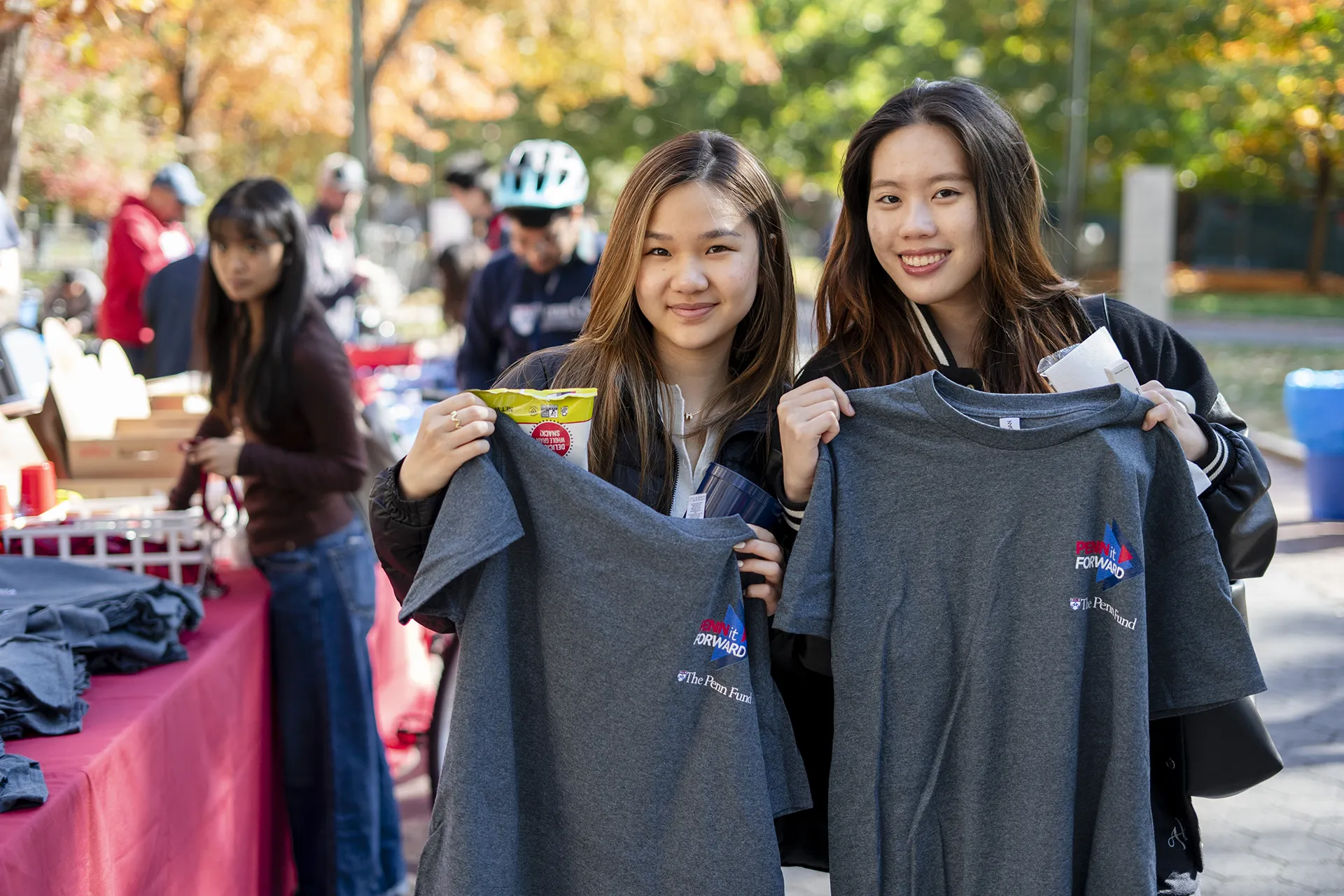 Two students smiling and holding gray event T-shirts at a Penn campus giveaway table.