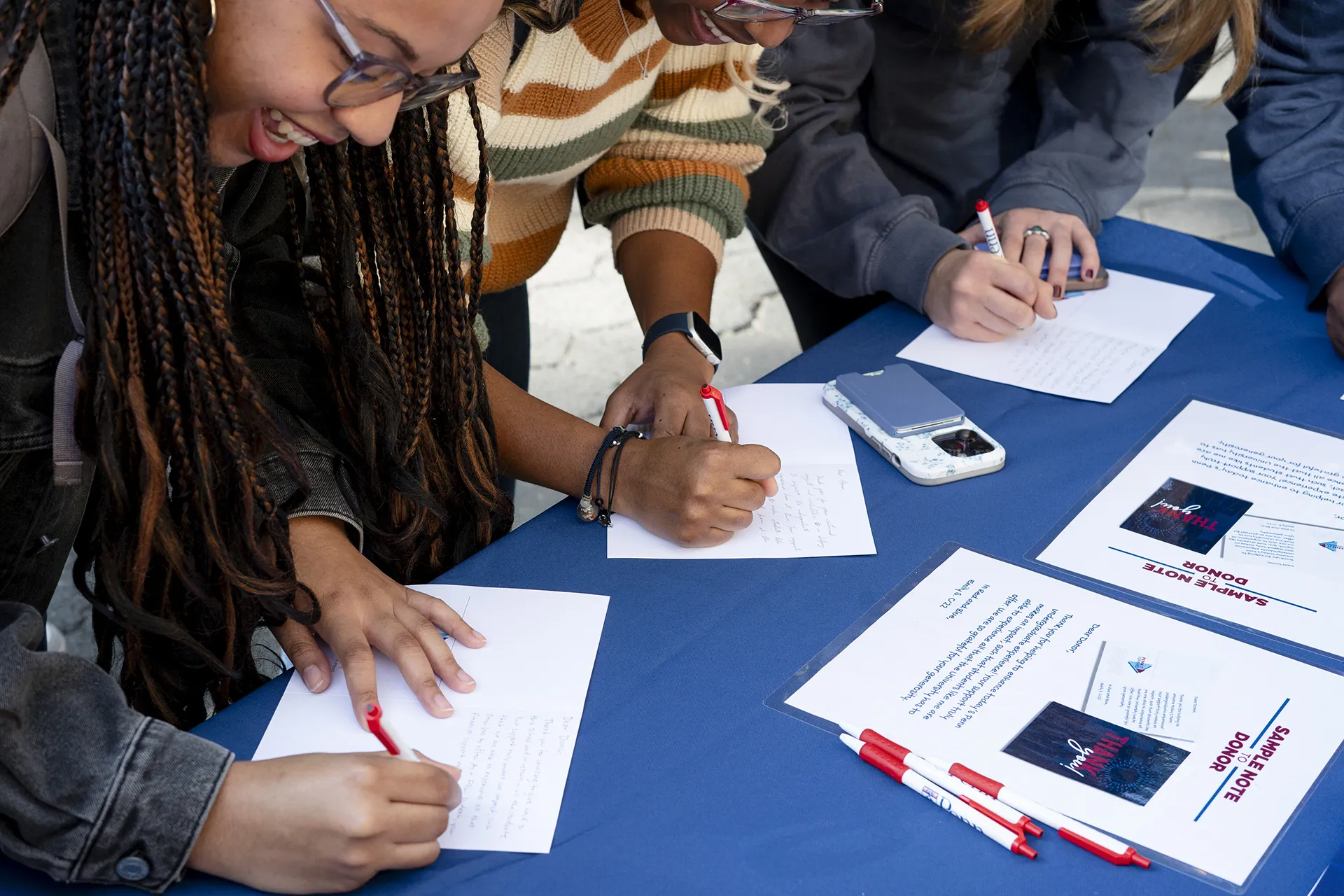 Students smiling and writing handwritten thank-you notes to alumni donors during a campus activity.